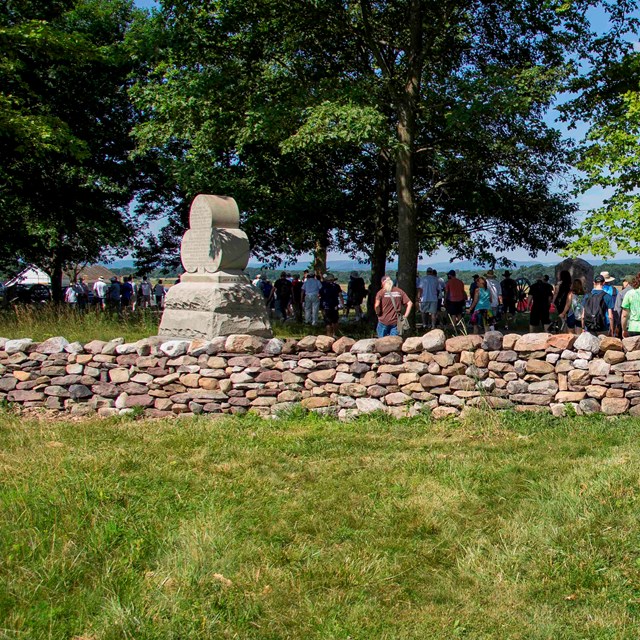 A large of visitors walk past a monument and a stone wall. Green grass is in front of the wall.