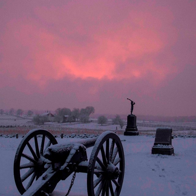 Snow covers the battlefield, a cannon and two monuments are in the center and pink and purple clouds