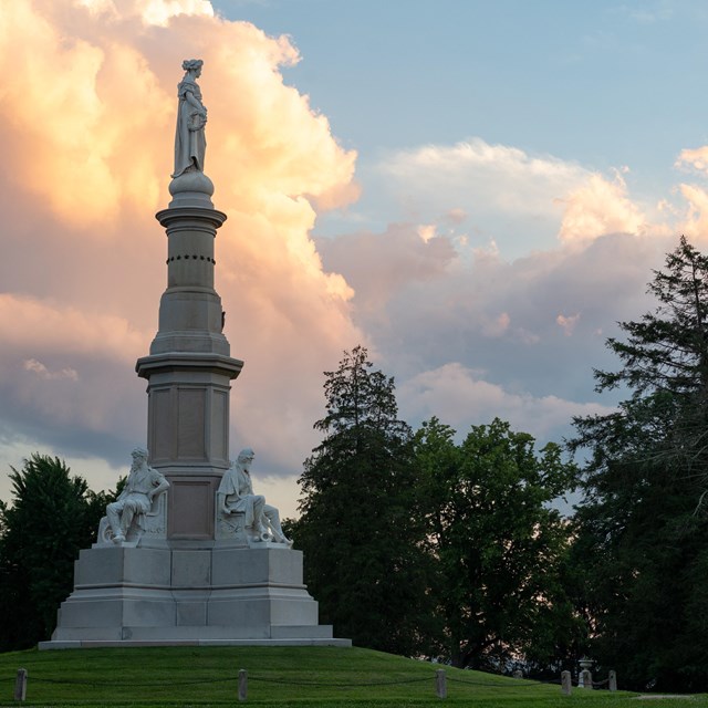 A stone monument with a woman on top surrounded by tress and clouds