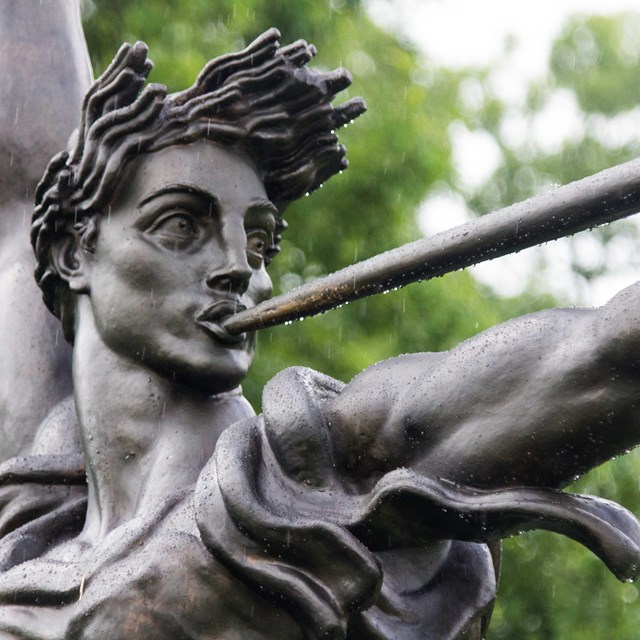 A dark bronze statue of a female figure is seen blowing a horn with a long tube.