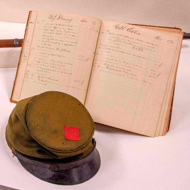 A faded green hat with red patch and black brim with an old notebook and Civil War rifle on a table.