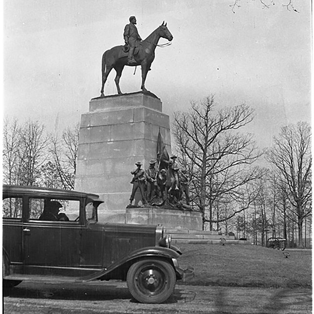 A black and white photo of a tall monument and bronze horse and rider on top. An old car is in front
