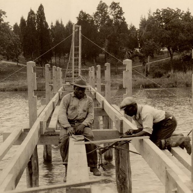 old photograph of two black men building a bridge over water.