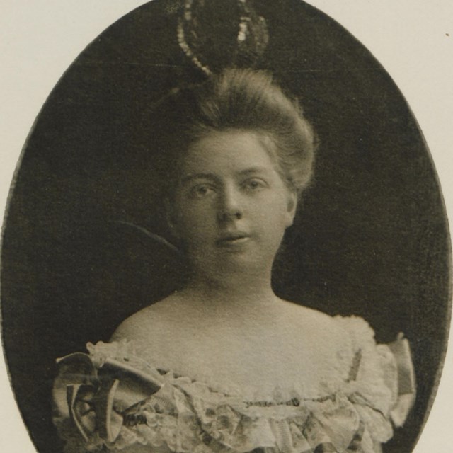 A black and white photo of a woman with a strapless dress with an elaborate hairdo and hat.