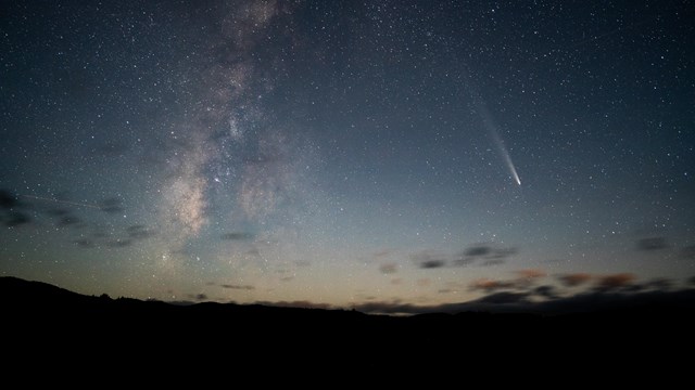 A bright comet with a glowing tail streaks across the night sky beside the hazy band of the Milky Wa