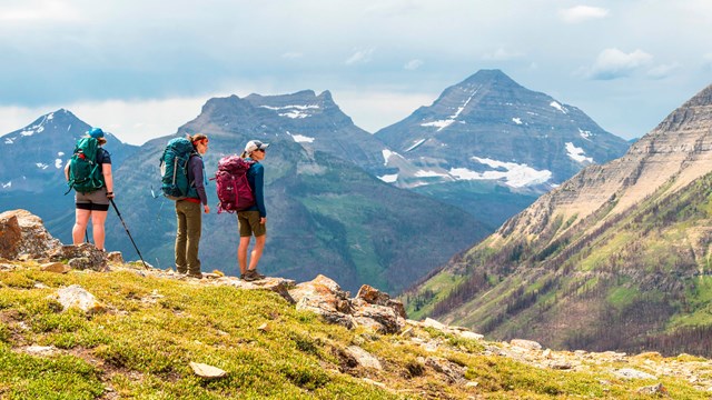 Three hikers stand on a clearing of a mountain looking out at a mountain range.