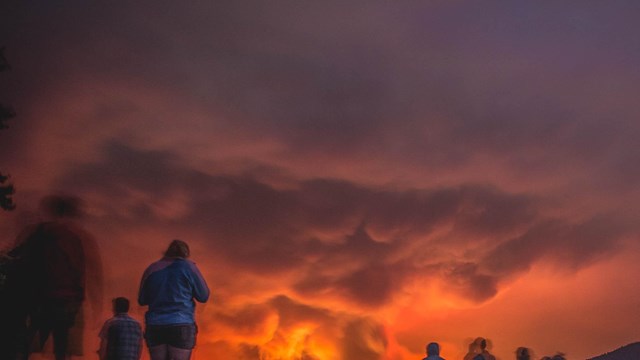 People stand on a beach and watch a wildfire. 