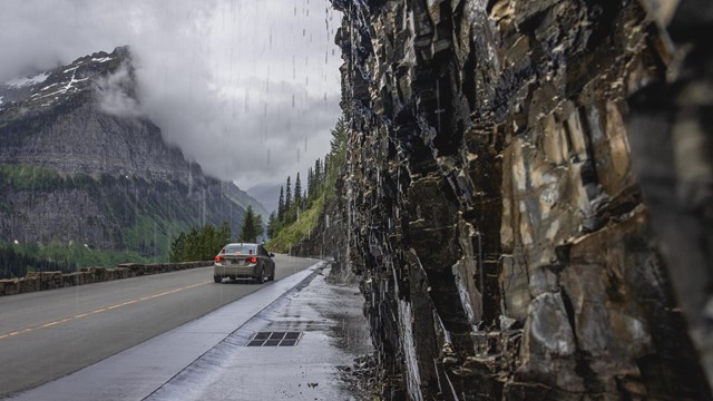 A vehicle drives on a narrow, alpine road. On the side of the road, water seeps down a wall of rock.