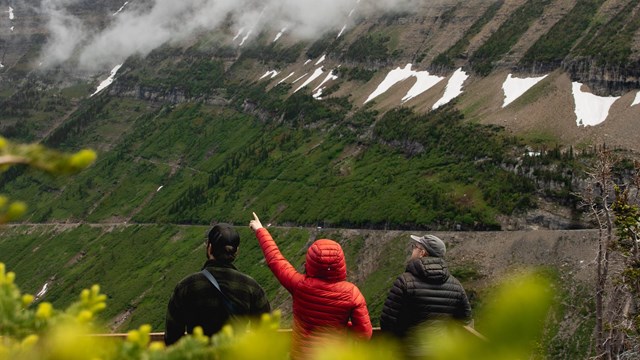 Three visitors stand at an overlook viewing mountains. The one in the middle points upwards.