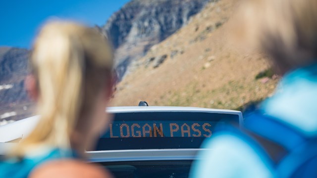 Two blurry people frame the image of the top of a shuttle bus that says, "Logan Pass." 