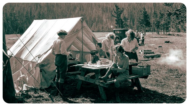 Group of people sit by a tent. Trees in the background. 