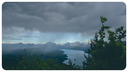 A bolt of sunlight shines through cloudy and hazy skies, hitting mountains and a lake below.