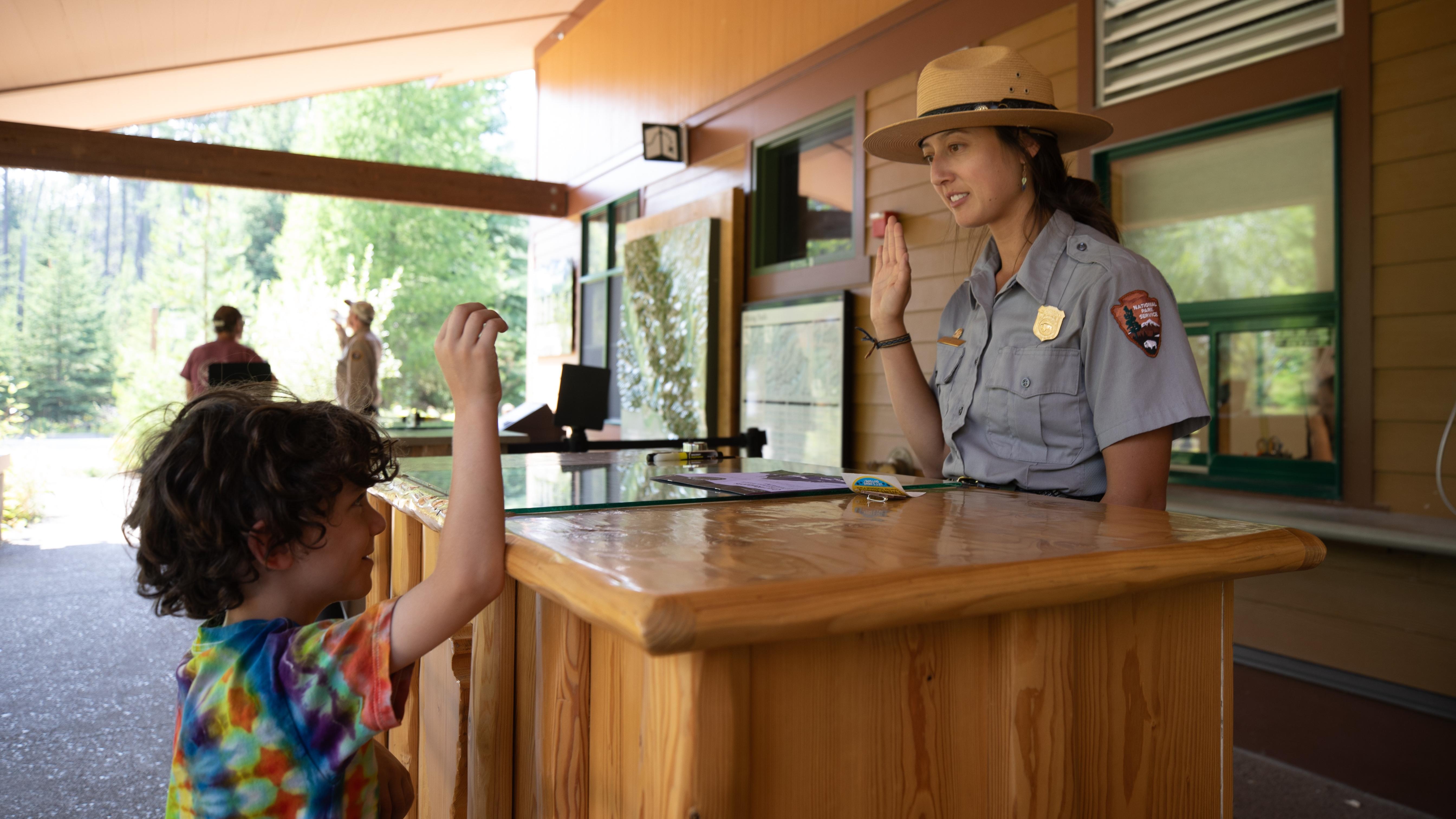 A young boy in a tie-dye shirt raises his right hand while looking at a park ranger doing the same.