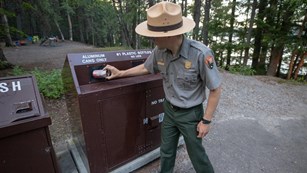 A park ranger deposits a soda can into a recycling bin with a forest in the background. 