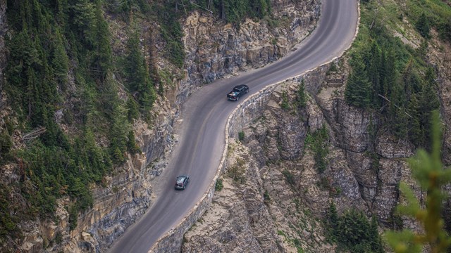 Two cars drive along a cliff's edge narrow road with trees all around. 
