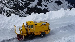 A large plow truck removes snow from a high alpine road with a vast landscape beyond and cloudy sky.