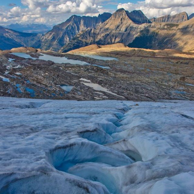 A view of glacial ice carved by water with a view of rocky mountains in the background.