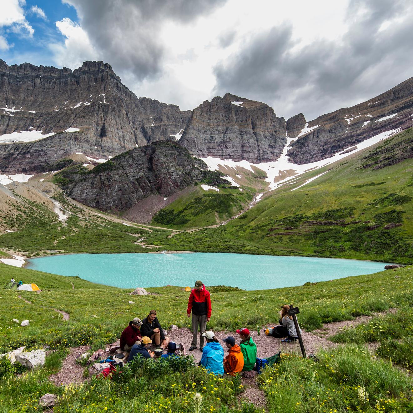 Visitors prepare a meal at a lakeside wilderness campsite near a mountain.