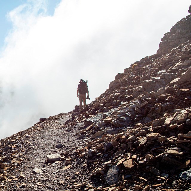 A person standing on a mountain trail with clouds behind. 