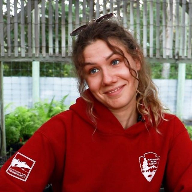 A woman in a red sweatshirt sits at a picnic table with her hands out looking confused and amused.