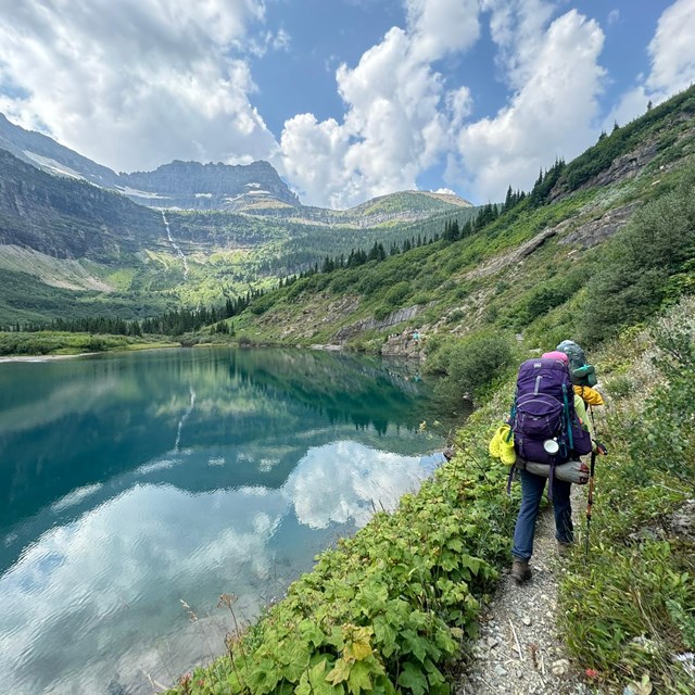 Two hikers with large backpacks walk down a trail that follows the edge of a turquoise lake.