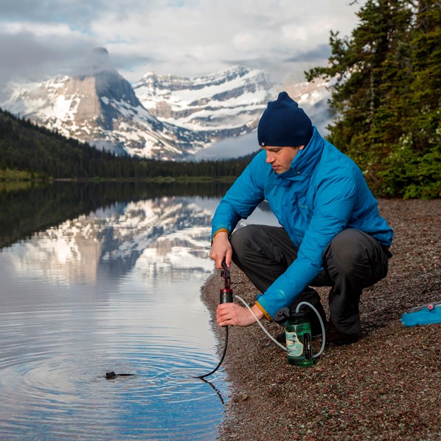 A man with hat and blue jacket uses a water filter to pump water out of a large lake.