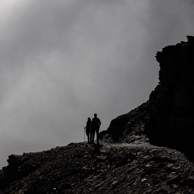 Two silhouetted hikers stand on top of a rocky mountain facing a wall of clouds. 