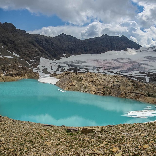 A bright lake surrounded by gravel, snow, and ice in the mountains. 