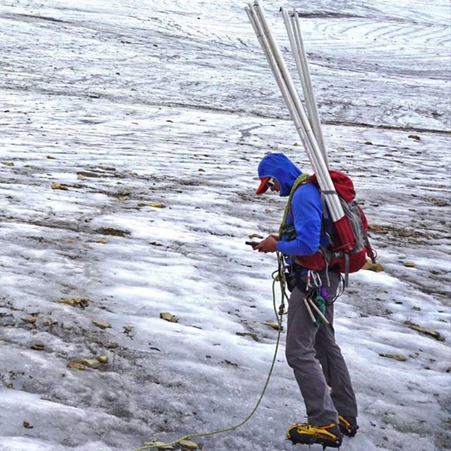 A researcher with a backpack holding long, plastic stakes stands on vast glacial ice.