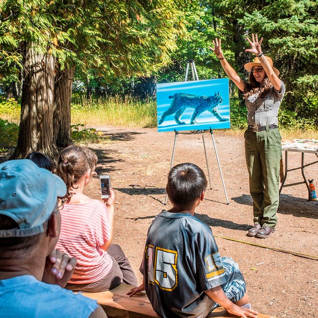 Ranger in front of easel throws her hands in the air while giving an outdoor presentation