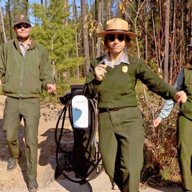 A park ranger dances in front of an electric vehicle charger on a sidewalk.