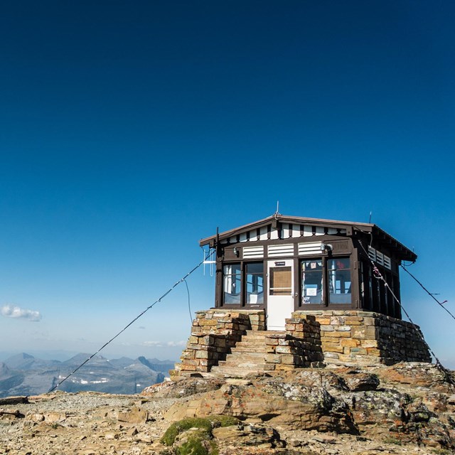 A small building with a stone foundation perches on a mountaintop, with snowy peaks beyond.