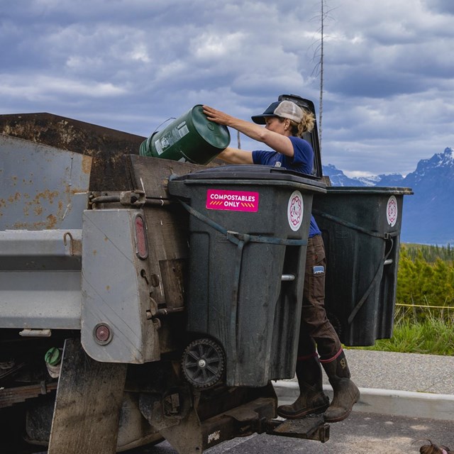 A ranger holds a green bucket in front of a composting truck with mountains in the background.