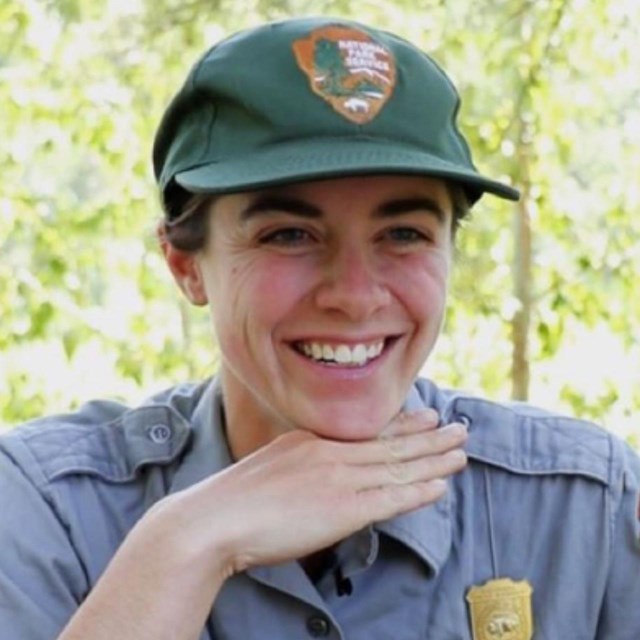 A ranger in a green and gray uniform sits smiling at a picnic table with her hand under her chin.