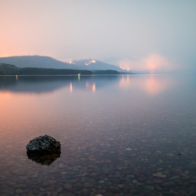 A smoky evening scene, with fire glowing on the far shore of a lake. The water is glassy.