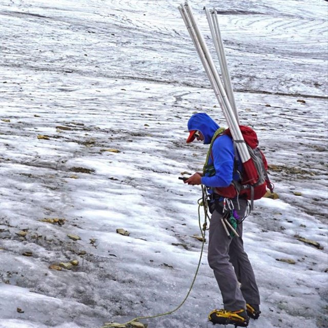 A researcher with a backpack holding long, plastic stakes stands on vast glacial ice.