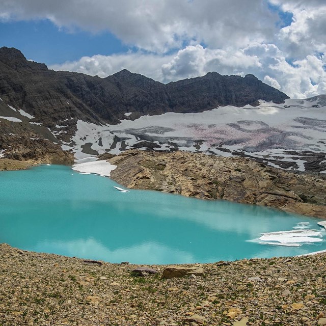 People walk around a bright lake with a glacier and mountains in the background. 