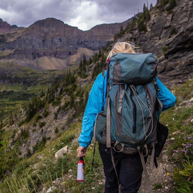 A backpacker walks on trail towards the mountains, holding a can of bear spray.