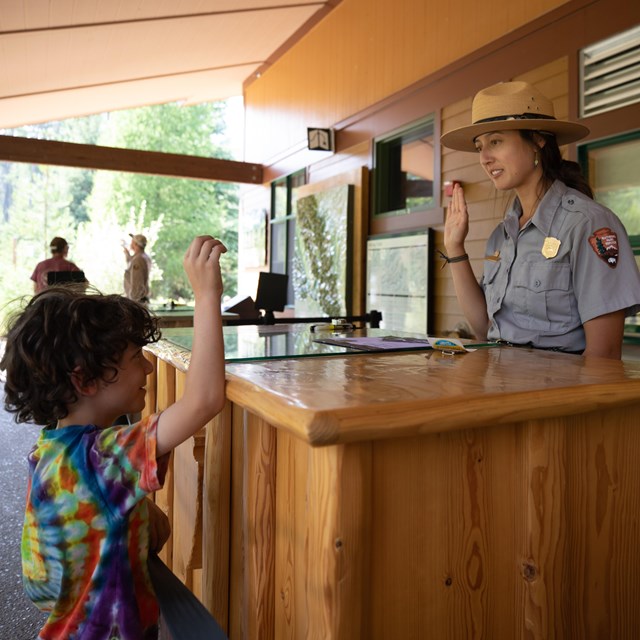 A young boy in a tie-dye shirt raises his right hand while looking at a park ranger doing the same.
