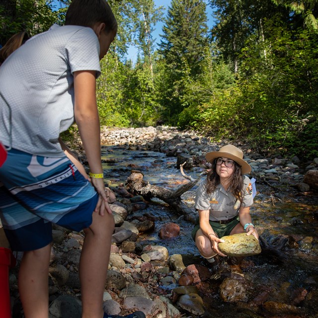 A ranger squats in a creek holding a large yellow rock while talking to a group of kids on the bank.