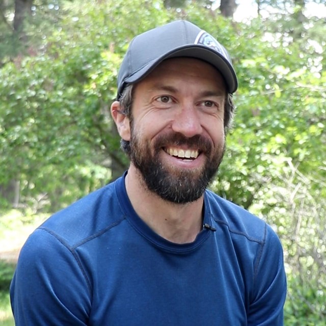 A man with a beard wearing a ball cap sits at a picnic table smiling at the camera.