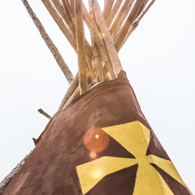 The top of a teepee lodge against a smoky sky with sun flaring through the poles.