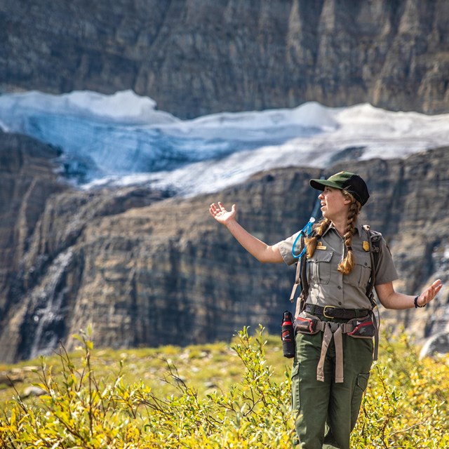 A park ranger stands with their arms out in front of a glacier. 