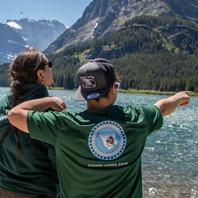 A youth points out at a lake with another youth in the background looking through a scope.