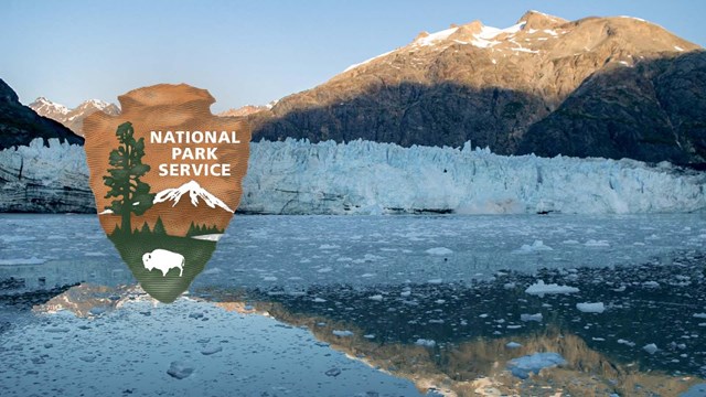 Calm water reflects a sun-topped mountain and glacier. NPS arrowhead logo overlaid.