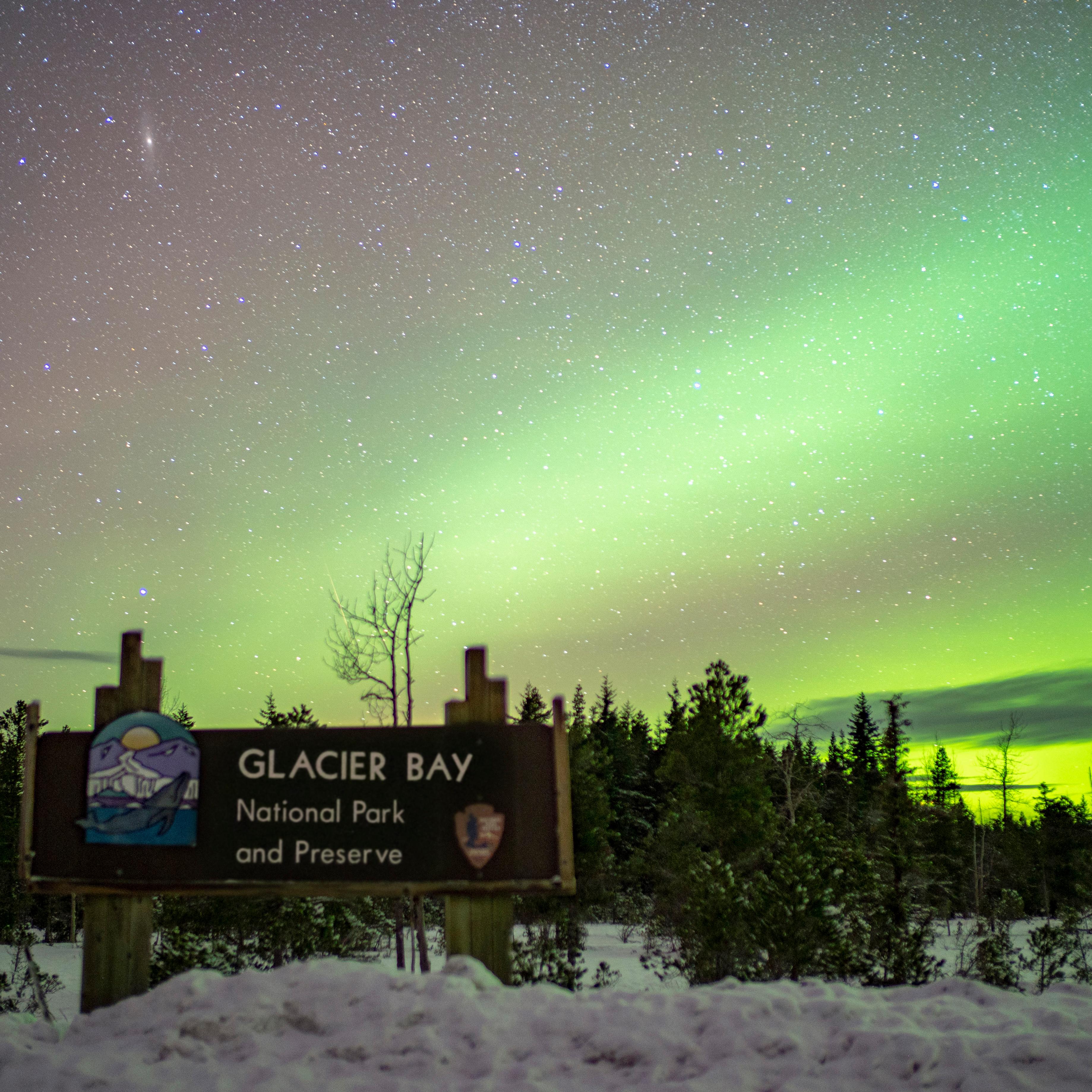 a green band of light in the night sky behind the snowy Glacier Bay entrance sign.