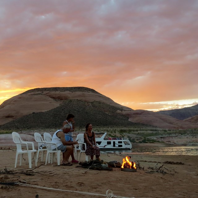 Two people sit in a gruop of lawn chairs around a campfire on a beach at sunset