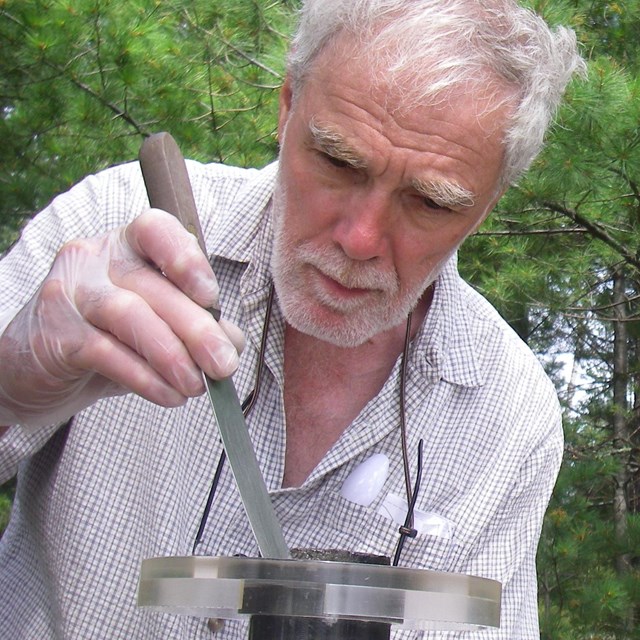 A gray-haired man carefully extracts a sample of lake-bottom sediment from a collection tube.