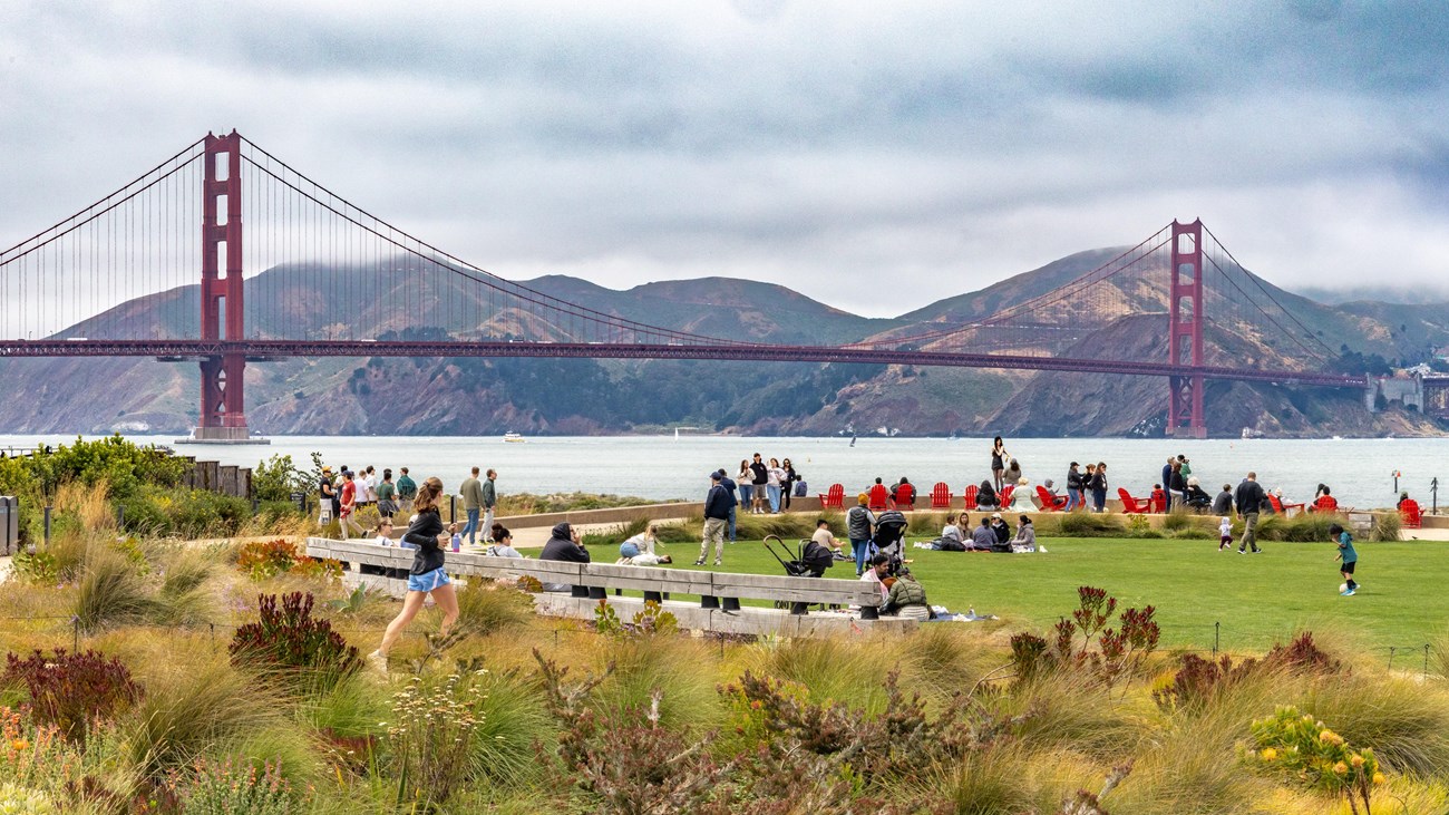 A photograph of people on benches and a lawn with the Golden Gate Bridge and mountains.