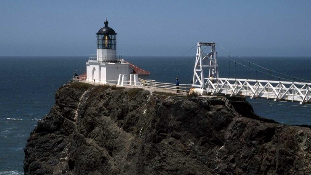 white lighthouse and suspension bridge atop cliff edge surrounded by ocean